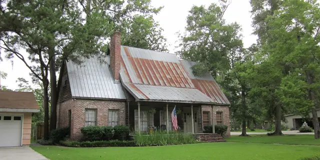 A small house with a shiny metal roof, a brick chimney and big shady trees