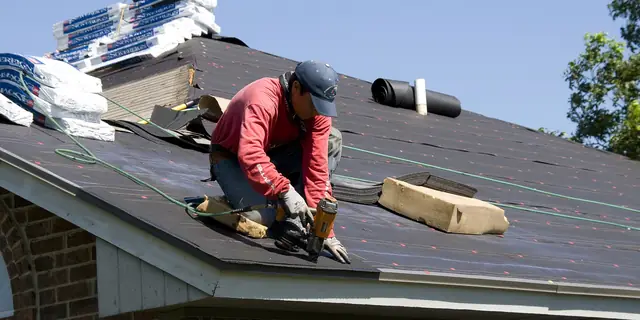 A roofer nailing down new shingles, with bundles of shingles beside him